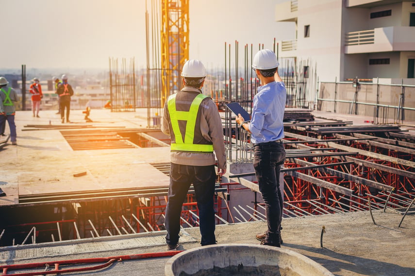 Two men in hard hats stand on top deck of commercial construction project reviewing the project while holding a tablet.