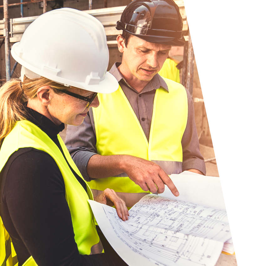 Man and woman both wearing hard hats and safety vest viewing construction blueprint together.