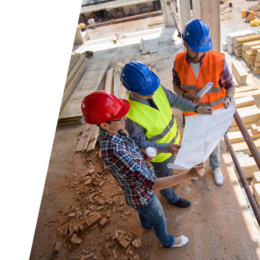 Three people wearing hard hats and safety vests stand on plywood floor surrounded by 2x4s while reviewing blueprints.