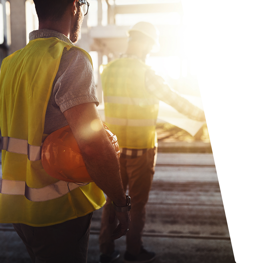 Two male construction workers wearing yellow safety vests evaluating blueprint with strong sunshine beam across the scene.